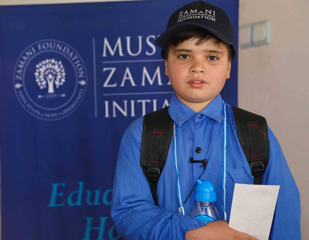 A young student in a Zamani Foundation cap in front of a Mustafa Zamani Initiative banner